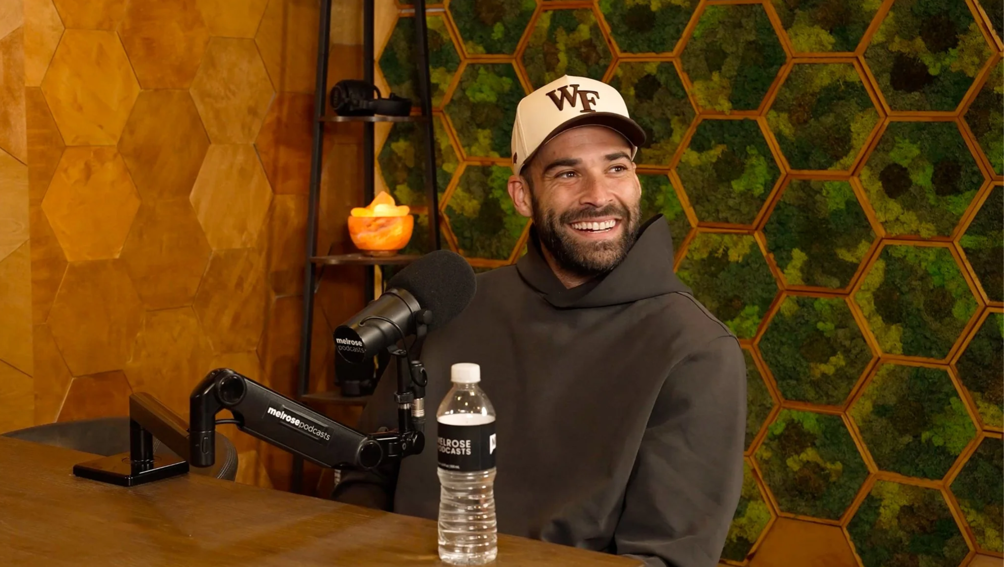A man with a beard smiling and wearing a white cap with "WF" on it, sitting at a table with a microphone in front of him, a water bottle, and a black hoodie. The background features a green moss wall with orange honeycomb frames and a small shelf with a bowl of what appears to be salt lamps.