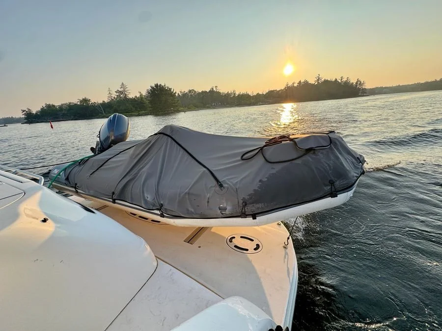 A boat with a covered motor and a covered dinghy on the deck sailing on a lake during sunset with trees in the background. Sea Ray 480 Sedan Bridge.