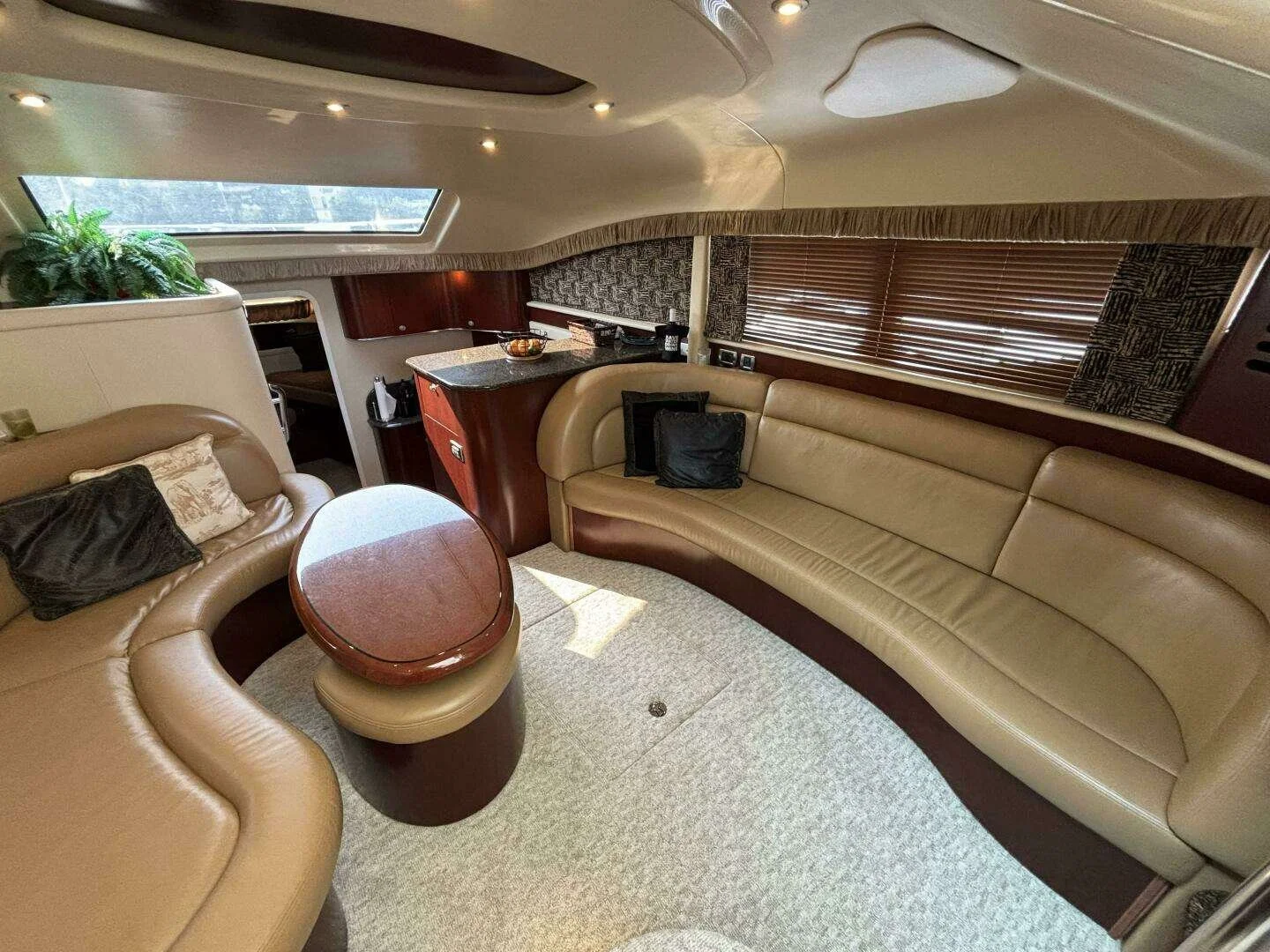 Interior of a luxury Sea Ray yacht with beige leather curved seating, a small oval wooden table, a kitchenette galley with wooden cabinets and granite countertop, and windows with wooden blinds.