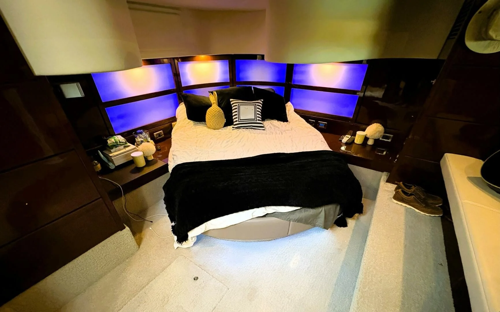 A cozy bedroom on a Sea Ray yacht, with a large bed, black and white pillows, plush toys, and blue-lit panels behind the headboard. Nightstands with lamps, books, and a pair of shoes on the carpeted floor.