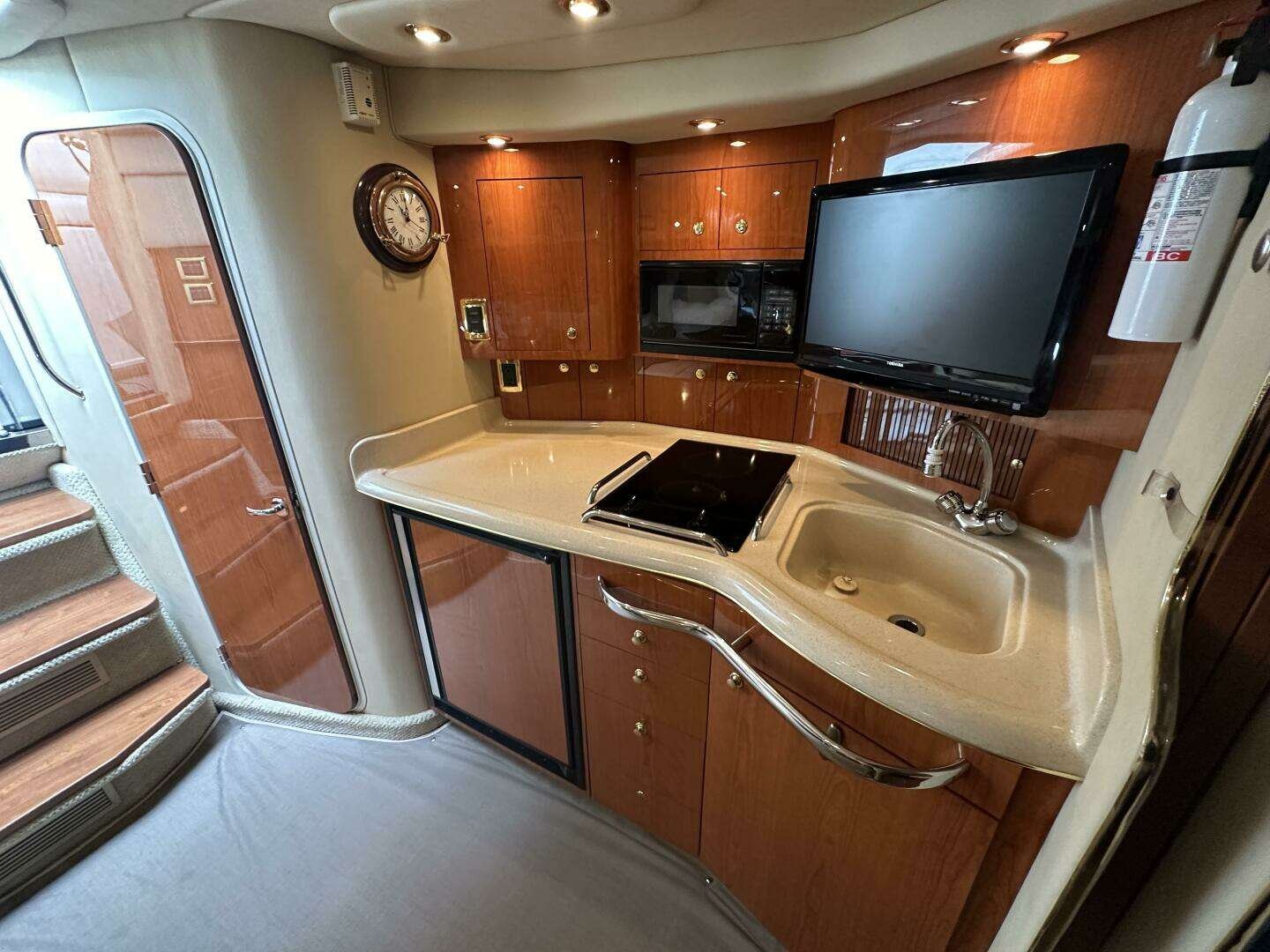 A boat's kitchen space with wooden cabinets, beige countertop, sink, black electric cooktop, microwave, wall clock, flat-screen TV, fire extinguisher, and staircase nearby.