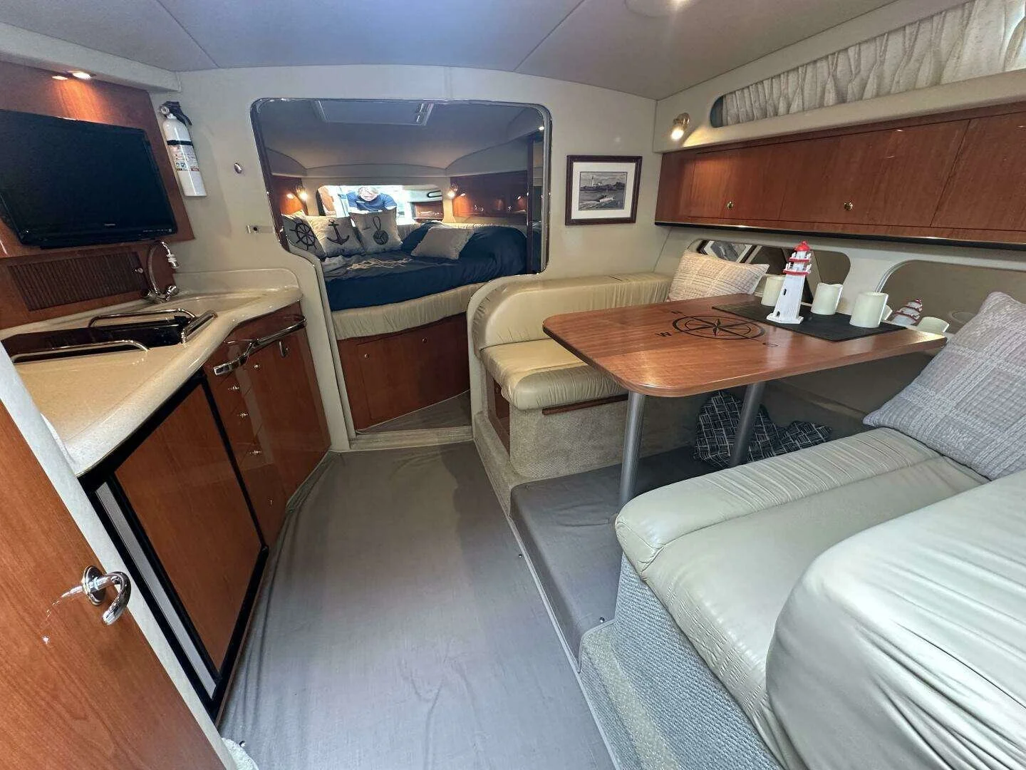 Interior of a yacht with a small kitchen area, a dining booth with nautical-themed decor, a television on the wall, and a bedroom visible through a doorway.