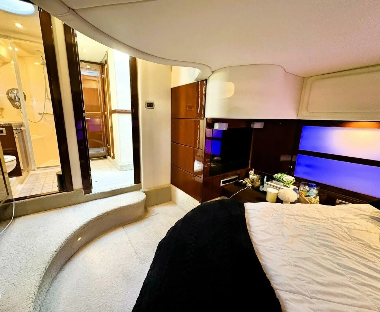 A Sea Ray yacht bedroom with a bed, a wooden entertainment center, and a TV with blue light. A view of a bathroom with a shower and toilet is visible through an open door. The room has beige carpet, and there are various items on the bedside table.