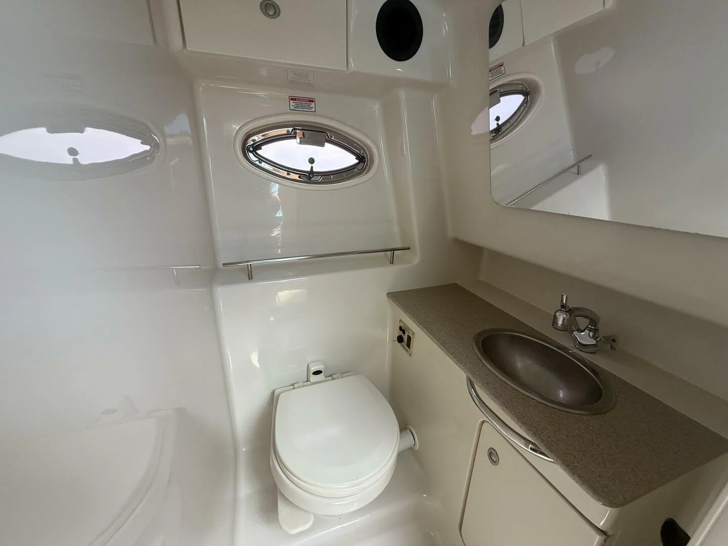 Marine head / bathroom with a toilet, a sink with a faucet, a mirror above the sink, and a porthole-style window.