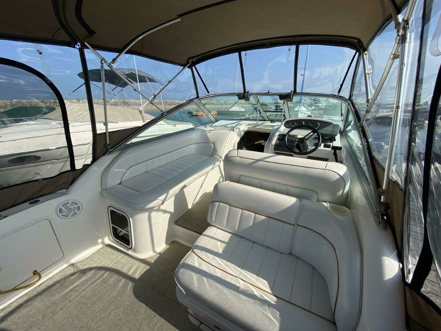 Interior of a boat featuring white cushioned seating, a steering wheel, and a dashboard with gauges, enclosed with clear plastic windows. Cockpit of Sea Ray 270 Sundancer.