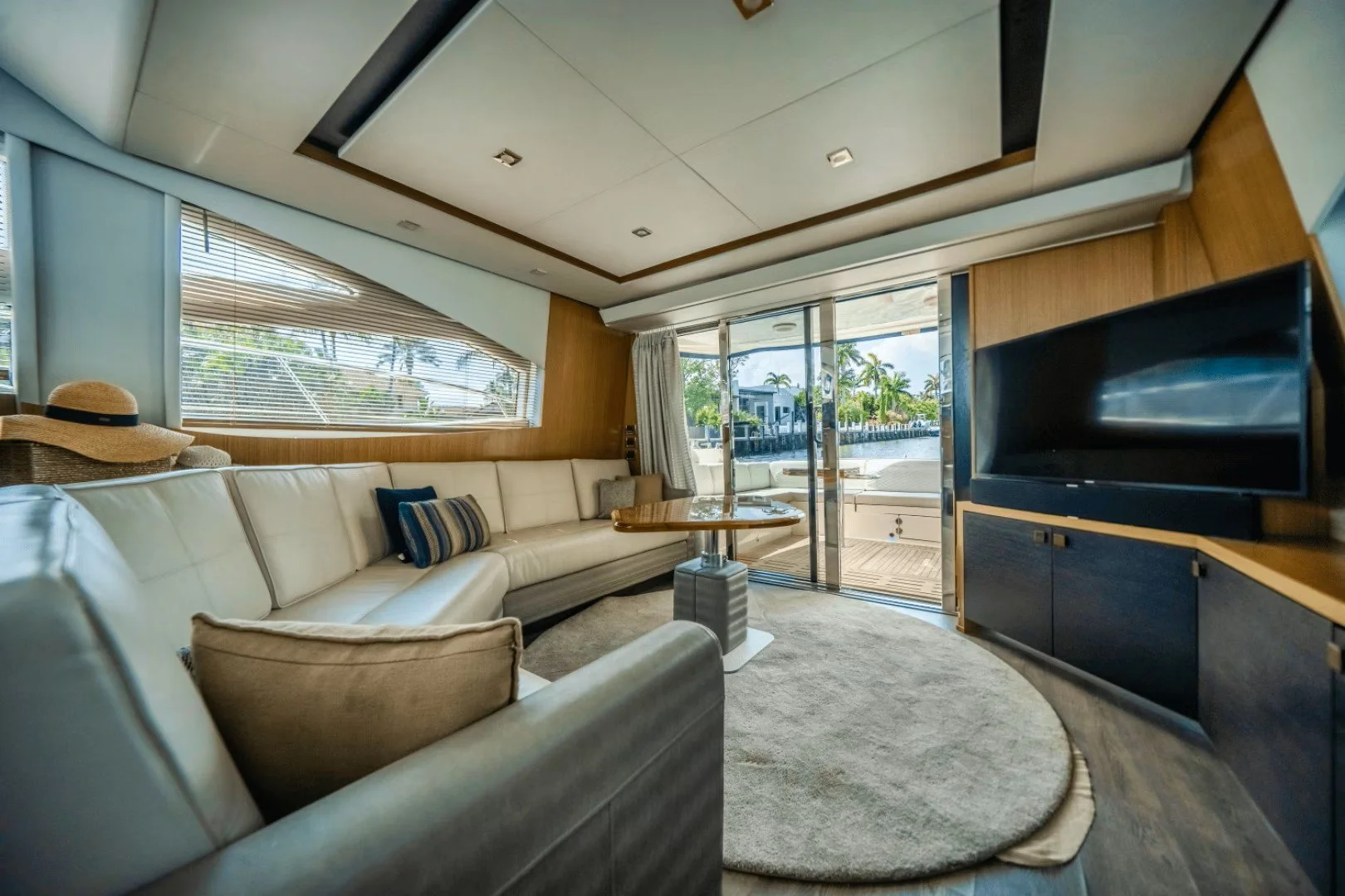 Interior of a modern Sea Ray yacht living room with large white sectional sofa, throw pillows, a wooden coffee table, a flat-screen TV, and large windows showing an outdoor patio and waterway with boats and palm trees.