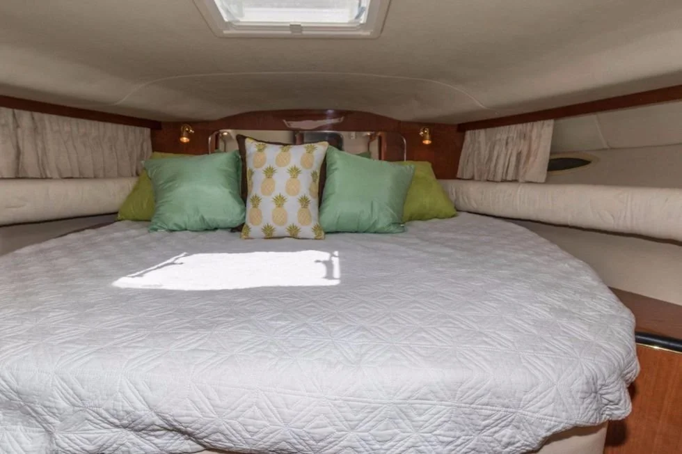 A neatly made bed with a white duvet and five pillows, including one with a pineapple print, inside a boat cabin.