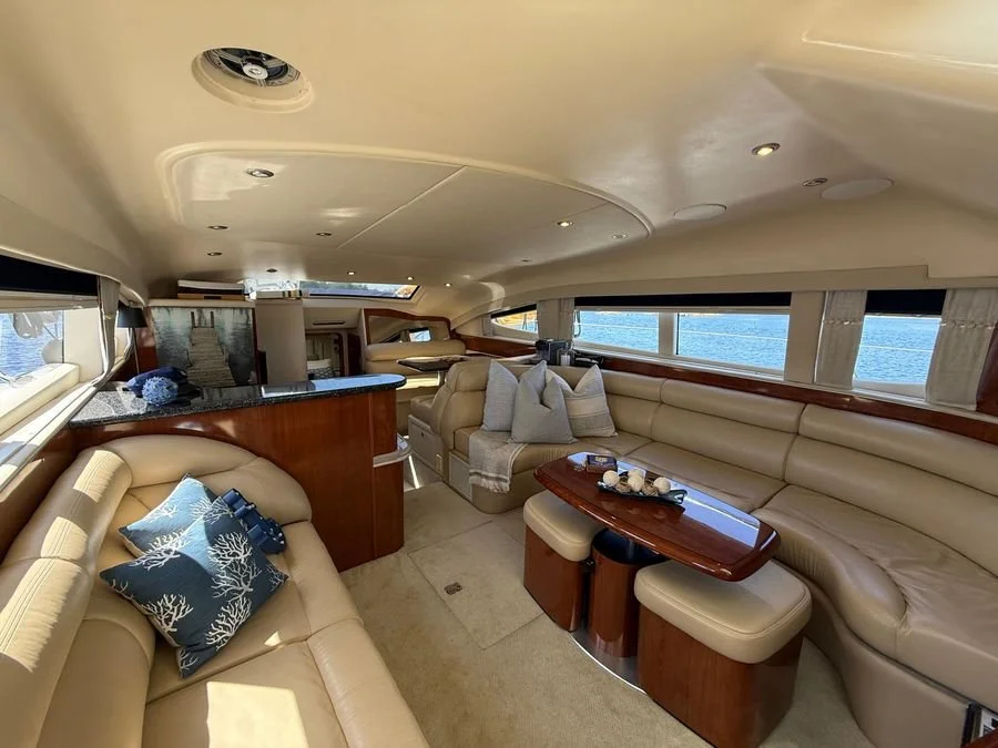 Interior of a luxury yacht with beige leather seating, decorative pillows, a wooden table, and windows showing water outside.