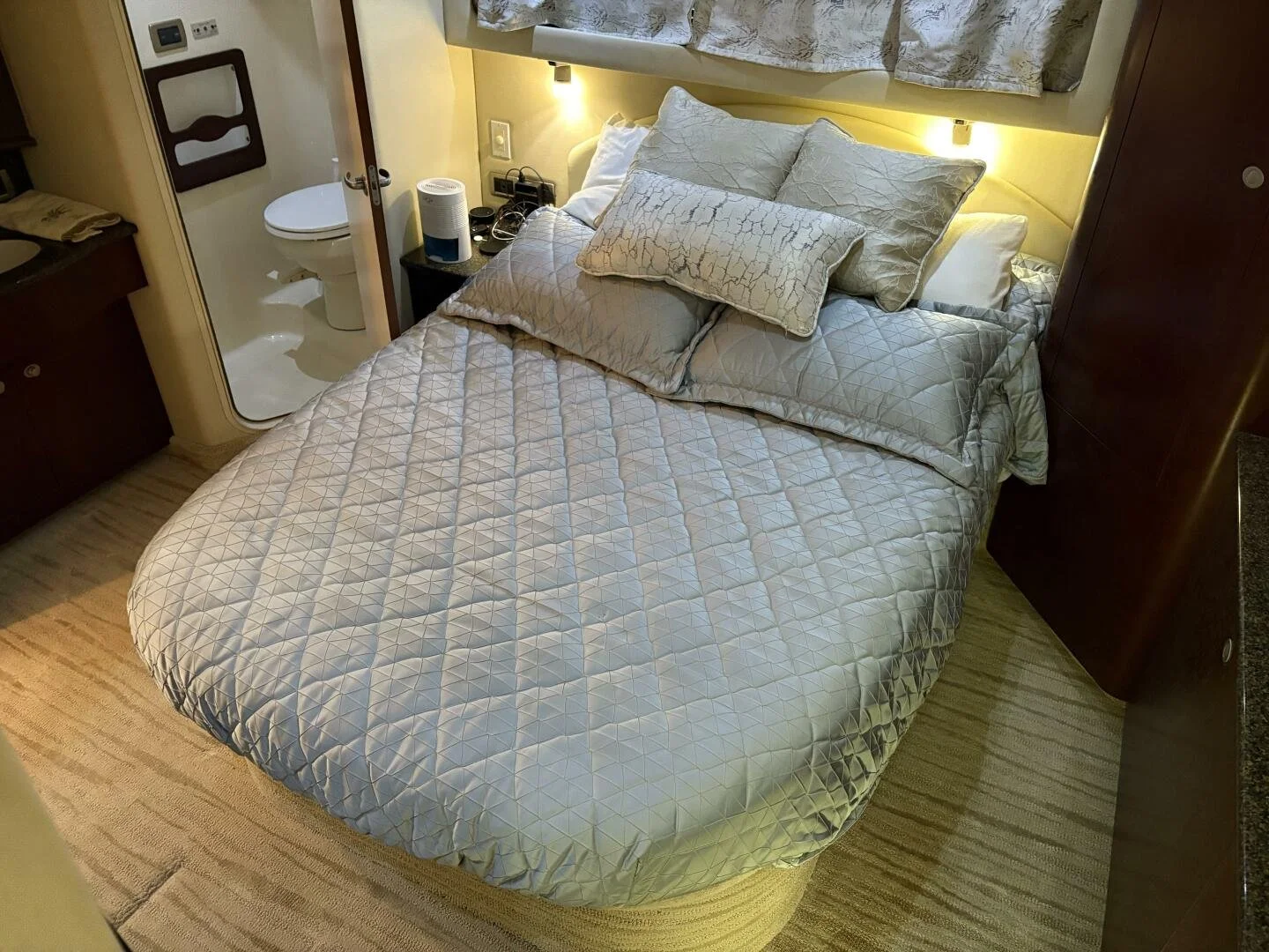 A neatly made bed with multiple white and silver patterned pillows on a Sea Ray yacht, with a visible bathroom through an open door to the left.