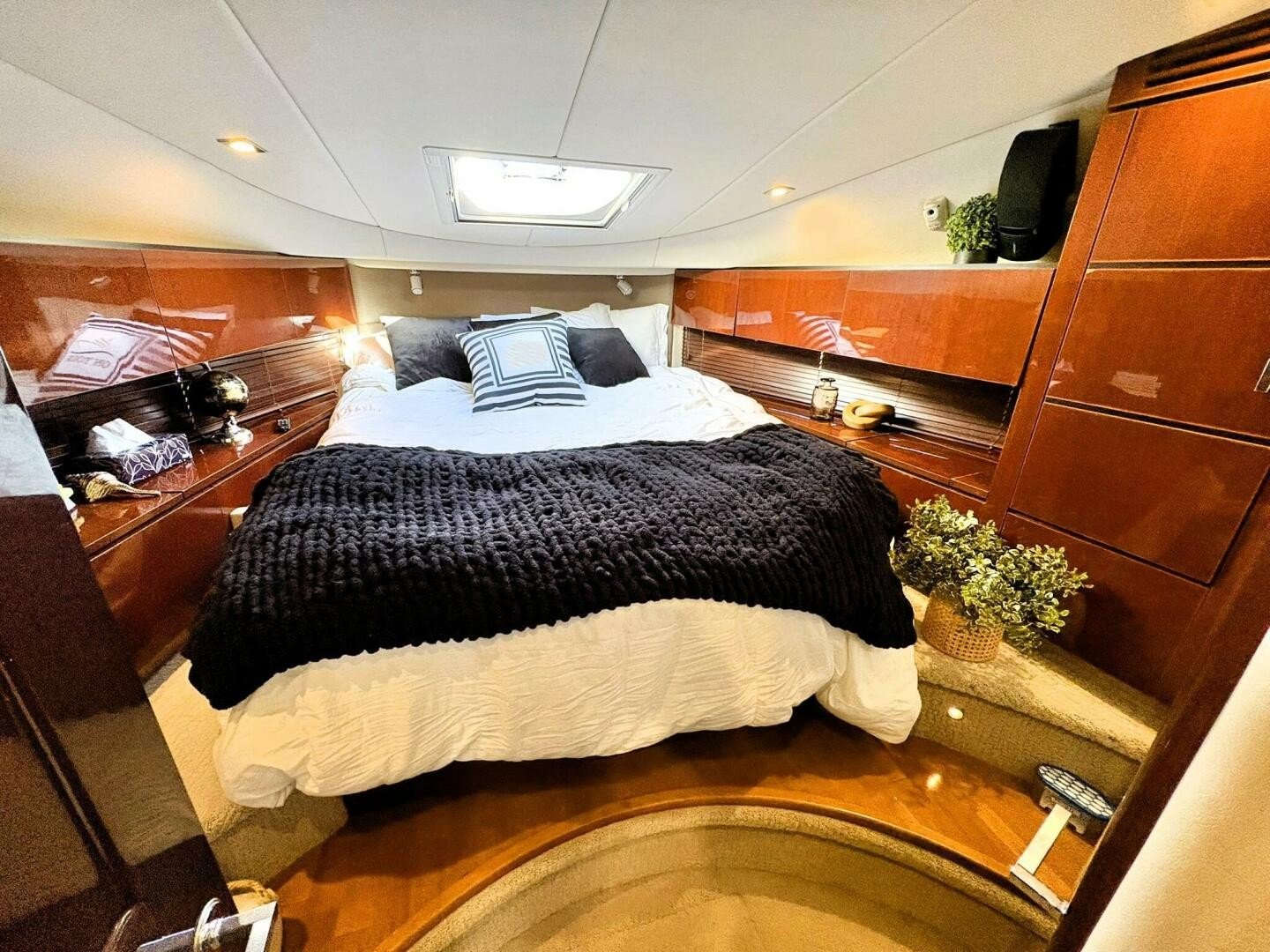 A cozy bedroom aboard Sea Ray yacht, with a wooden interior, a bed with white bedding and black accents, a small window above, and decorative items like plants, lamps, and a clock.