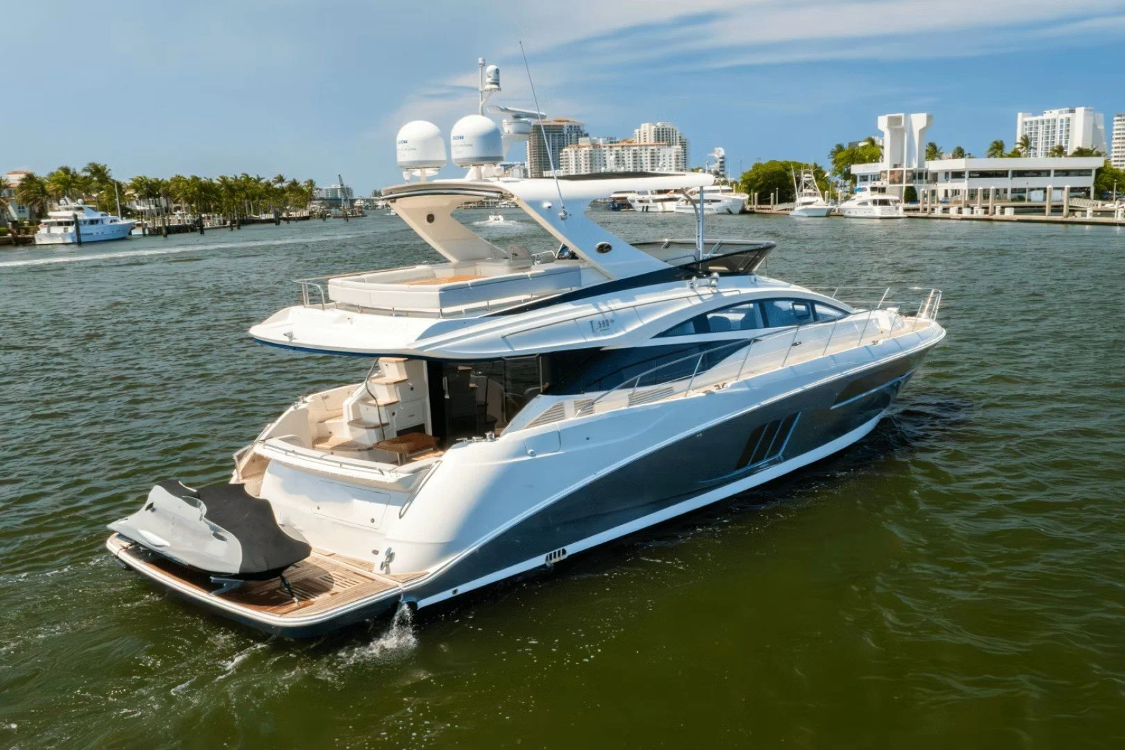 Luxury Sea Ray yacht cruising in a marina with high-rise buildings in the background and blue sky overhead.