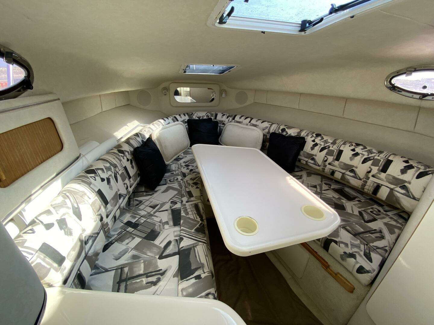 Interior of a boat cabin with a U-shaped seating area, patterned upholstery, a white table with cup holders, small windows, and a ceiling hatch. Sea Ray 270 Sundancer.