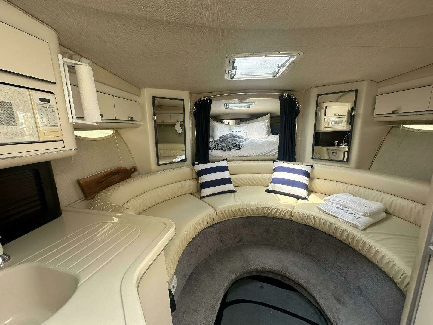 Interior of a Sea Ray sport boat, with a circular couch, striped pillows, a bed in the back, windows, and a microwave.