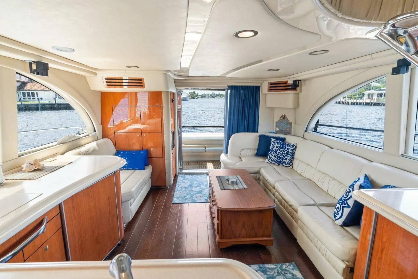 Interior of a boat with large windows, a white L-shaped sofa with blue patterned pillows, a wooden coffee table, and a kitchenette with wooden cabinets. Window view shows water and neighboring boats.