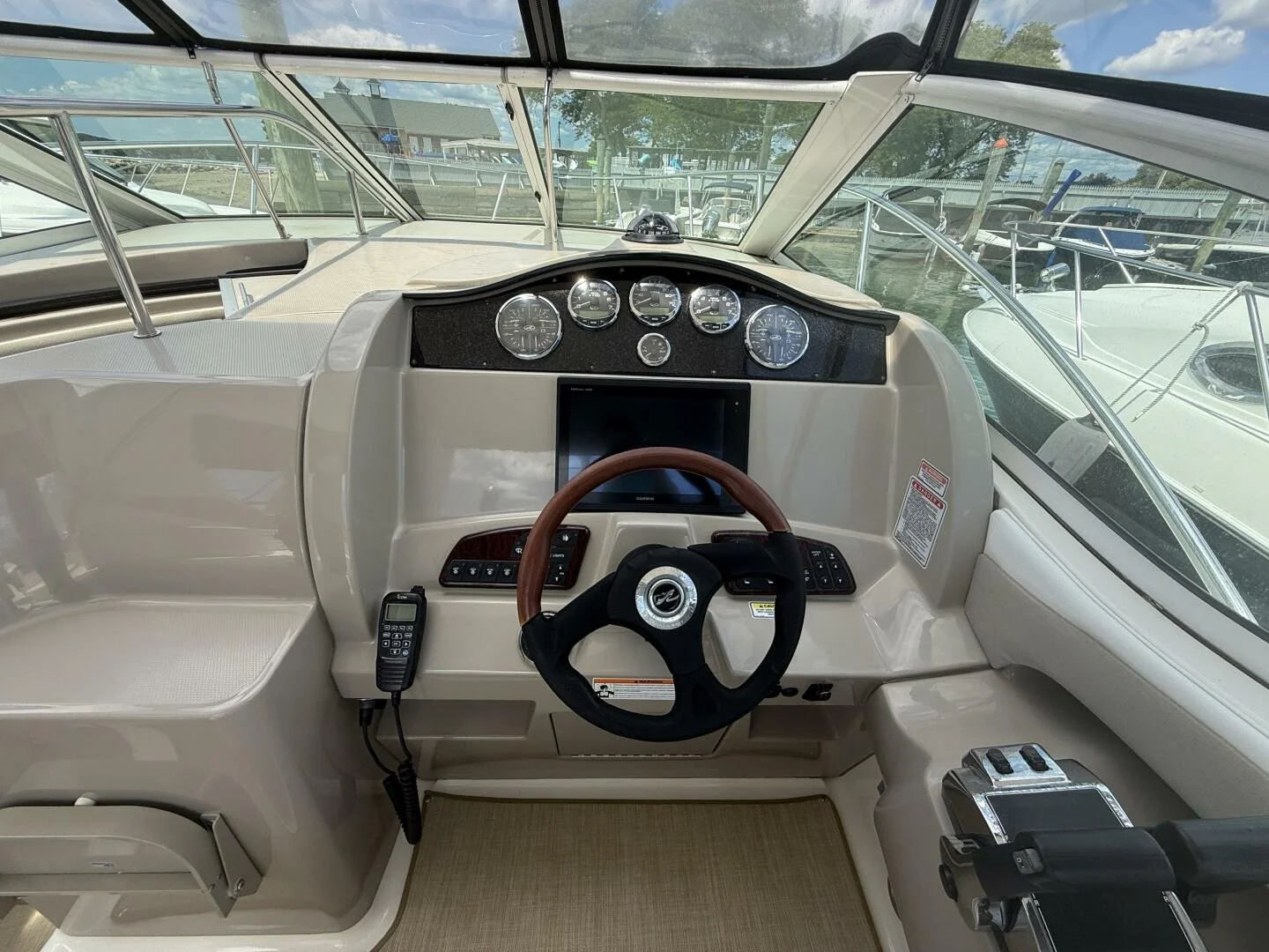 The interior dashboard of a boat with gauges, a touchscreen, steering wheel, communication device, and throttle controls, with windows showing a marina with other boats outside.