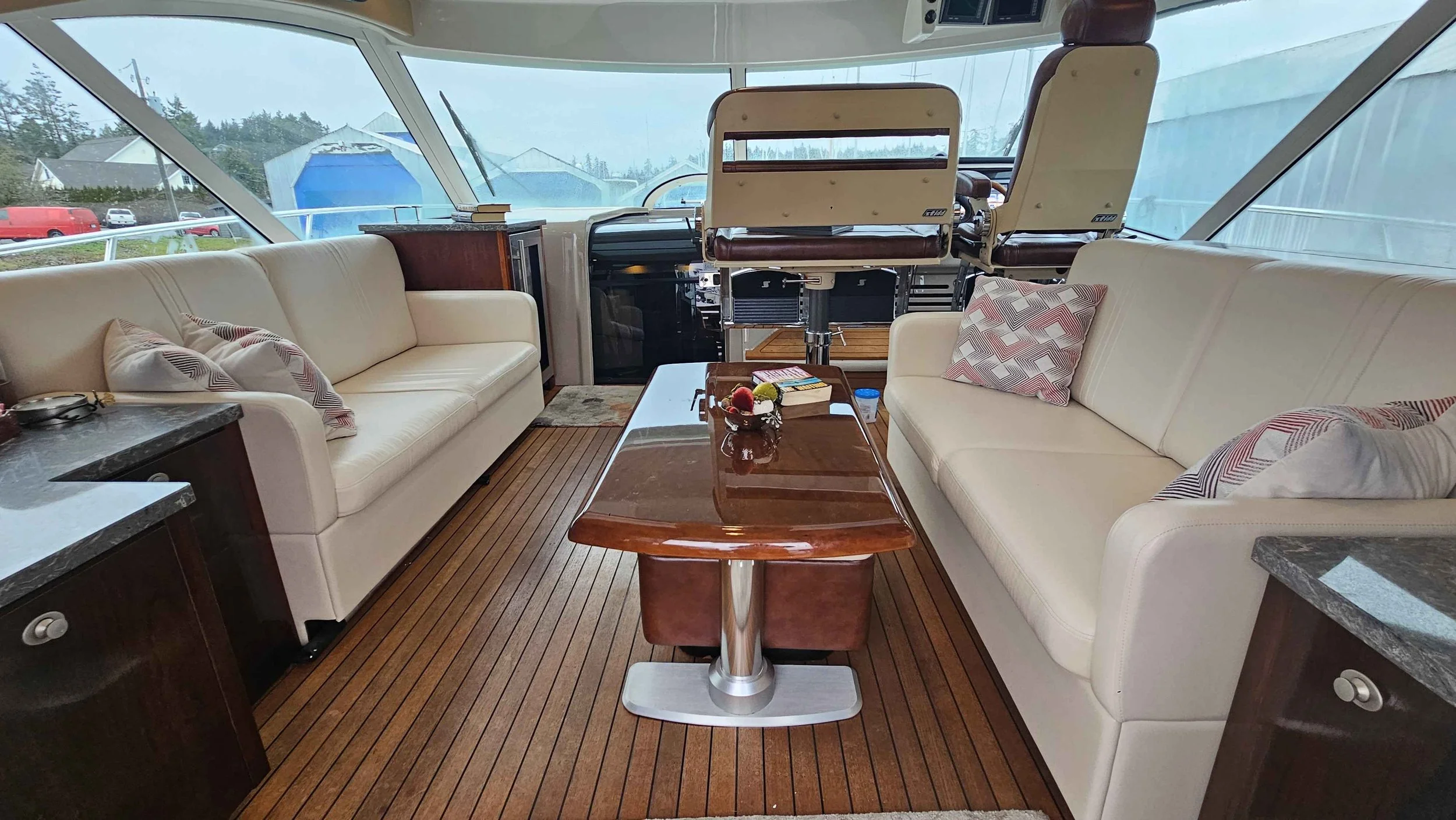 Interior of a boat with two white sofas on either side of a wooden table, a small countertop with a granite top on each side, and a view of the dashboard and windshield at the front.
