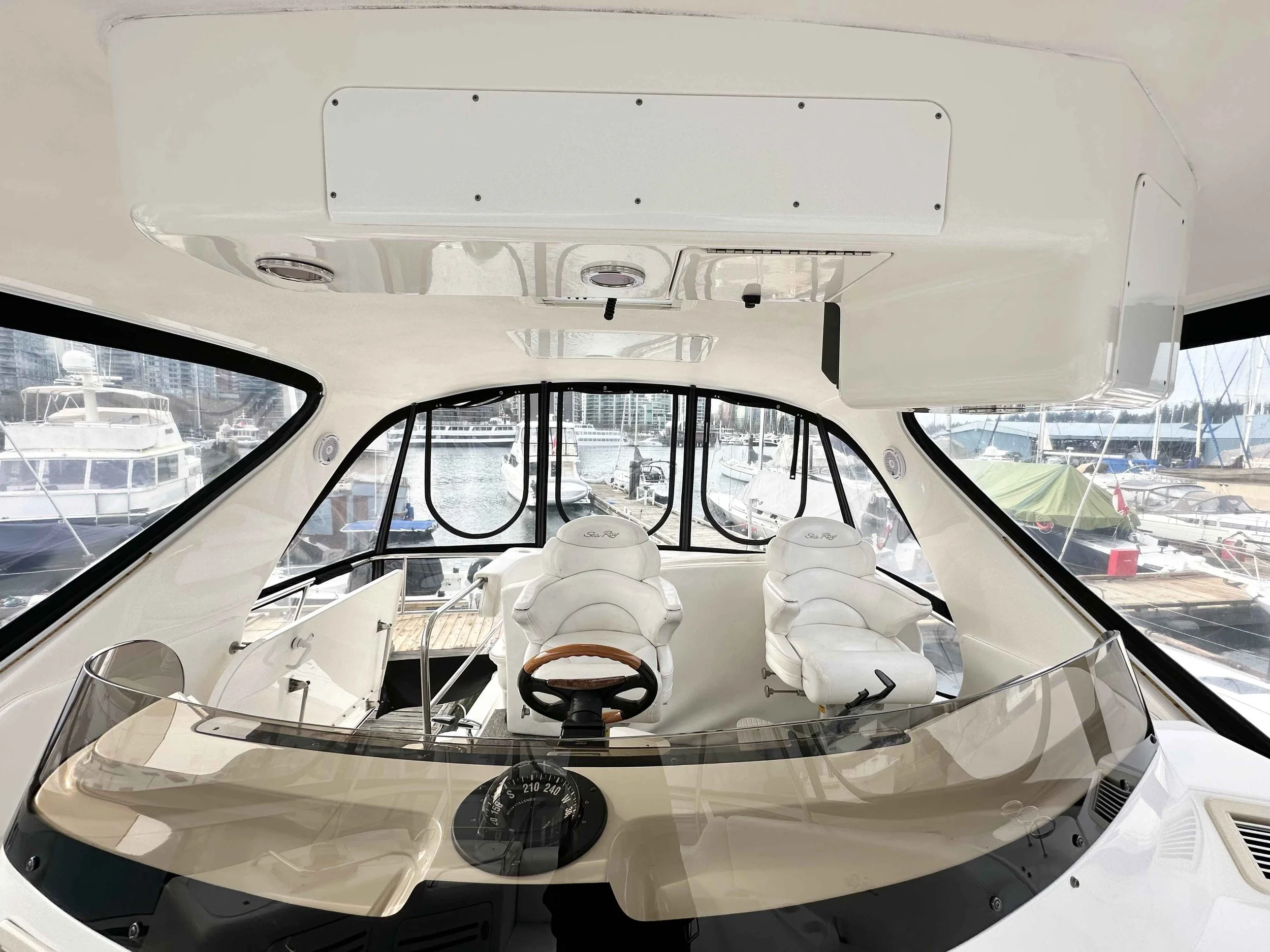 Interior of a yacht's steering area with two white captain chairs, a wooden steering wheel, and a compass on the dashboard, with large windows showing docked boats outside.