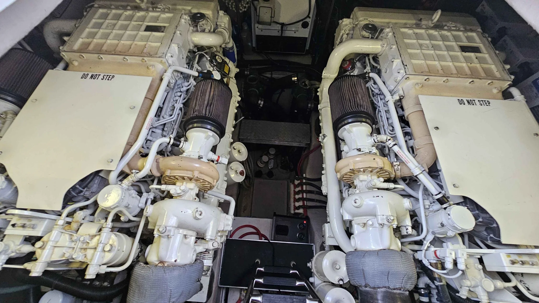 Sea Ray yacht engine room with two large, Cummins diesel engines labeled "Do Not Step." The engines have black air filters on top, various pipes, and machine, with black cabling and metal steps at the bottom of the image.