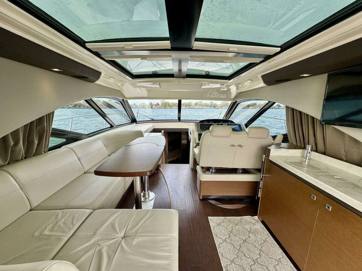 Interior view of a yacht's wheelhouse with cream seating, a wooden dining table, a navigation station, and a flat-screen TV, overlooking a body of water.