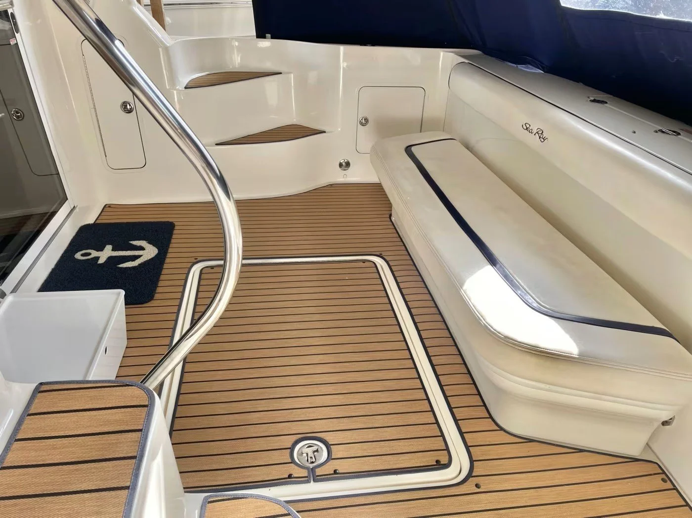 The image shows the deck area of a boat, featuring a beige cushioned bench, wooden steps, a black anchor rug, and custom wood-like flooring with a hatch cover. The boat's name, 'Sea Ray,' is visible on the side.
