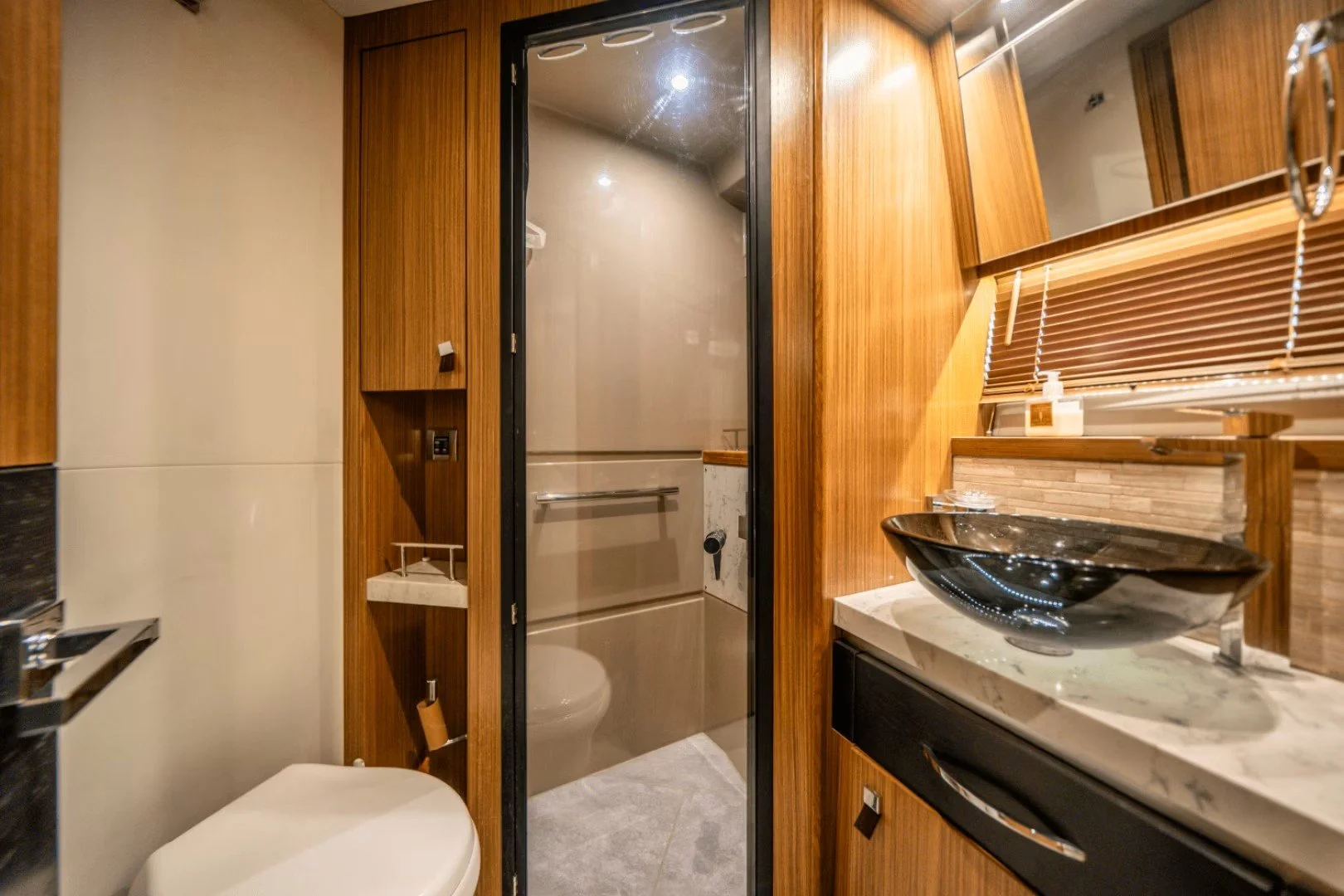 A yacht bathroom with cherry wood cabinetry, a toilet, a sink with a black vessel and a marble countertop, a mirror, and a small shower behind a clear glass door.
