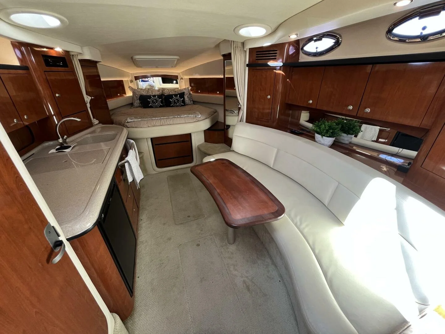 Interior of a luxury Sea Ray 340 Sundancer yacht with a beige curved sofa, a small wooden table, galley with a sink, and a sleeping area with a round bed and pillows. Wooden cabinets and portholes are also visible.