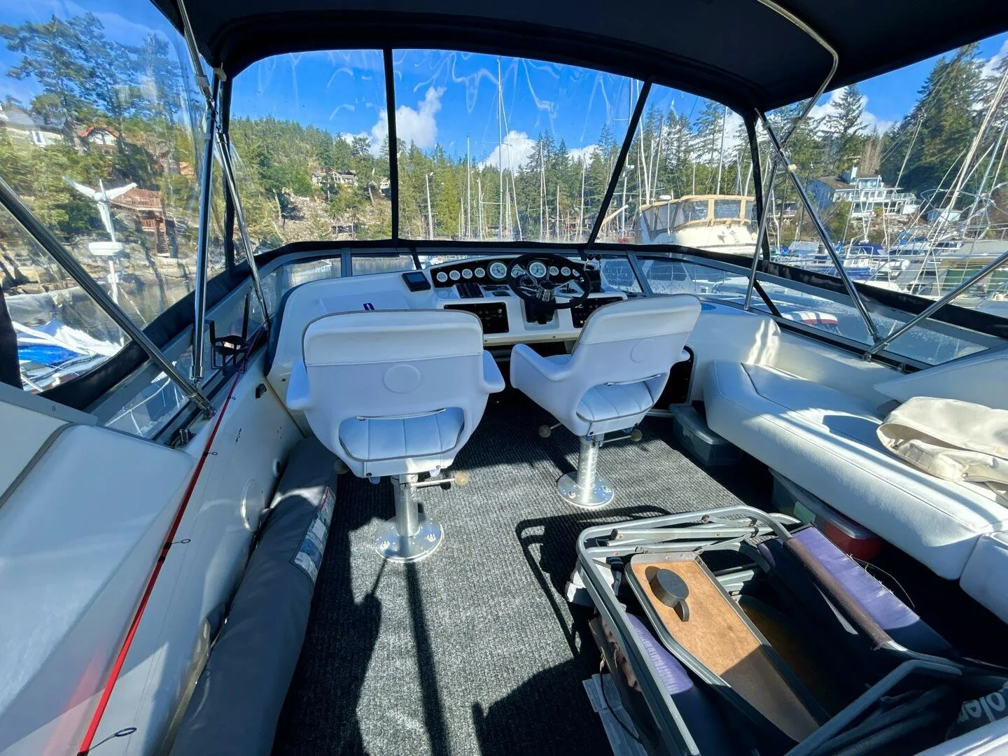 View of the interior of a boat with white chairs, dashboard, and steering wheel, docked at a marina with other boats and trees in the background on a sunny day.