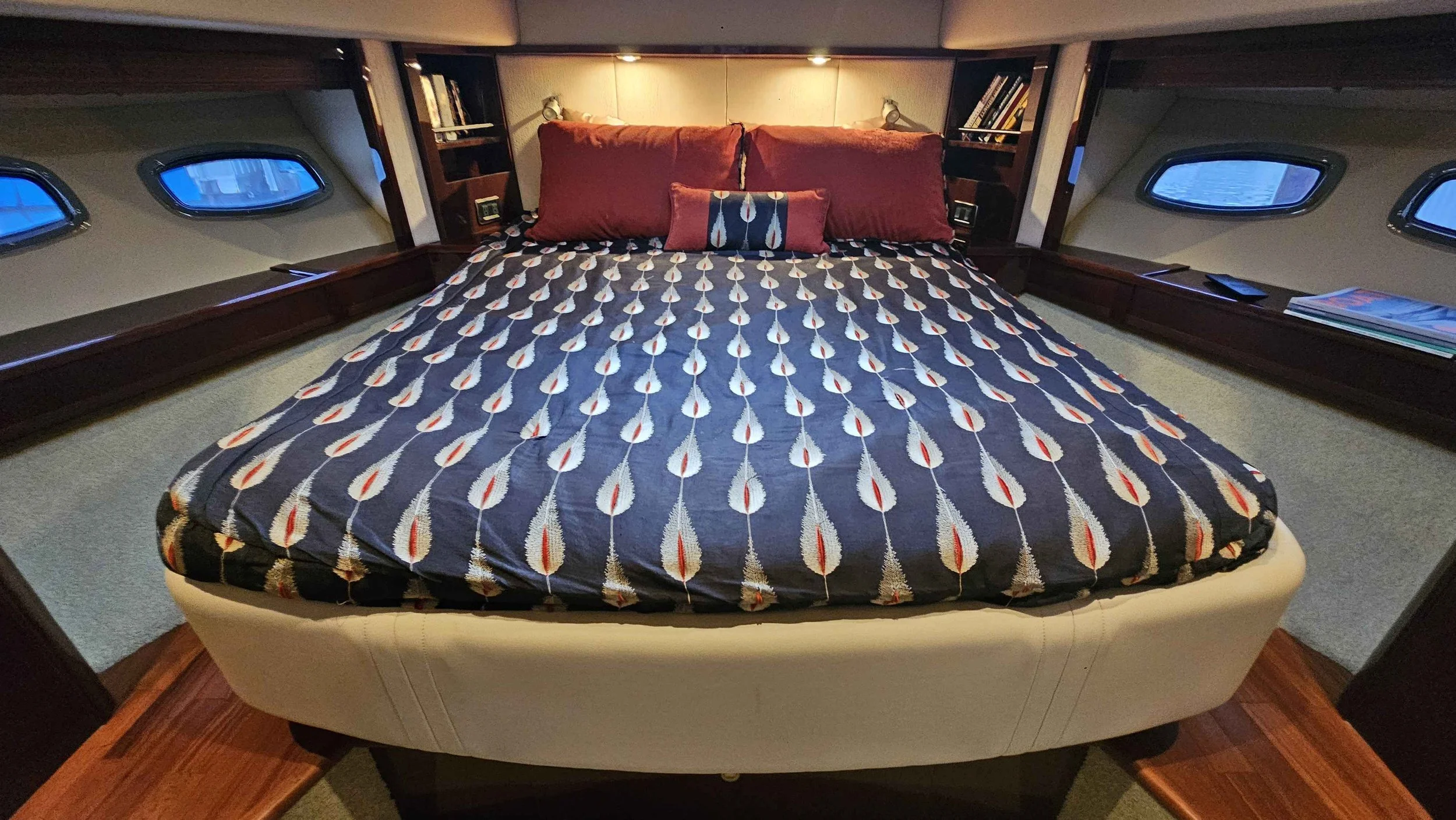A Sea Ray yacht's bedroom with a bed featuring a matching patterned bedspread and pillows, built-in side shelves, small windows, and reading lamps.