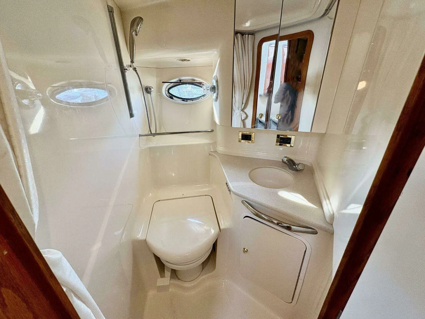 A white marine bathroom with a toilet, a sink with a curved countertop, a mirrored cabinet, a small porthole window, and a shower area. Sea Ray yacht.