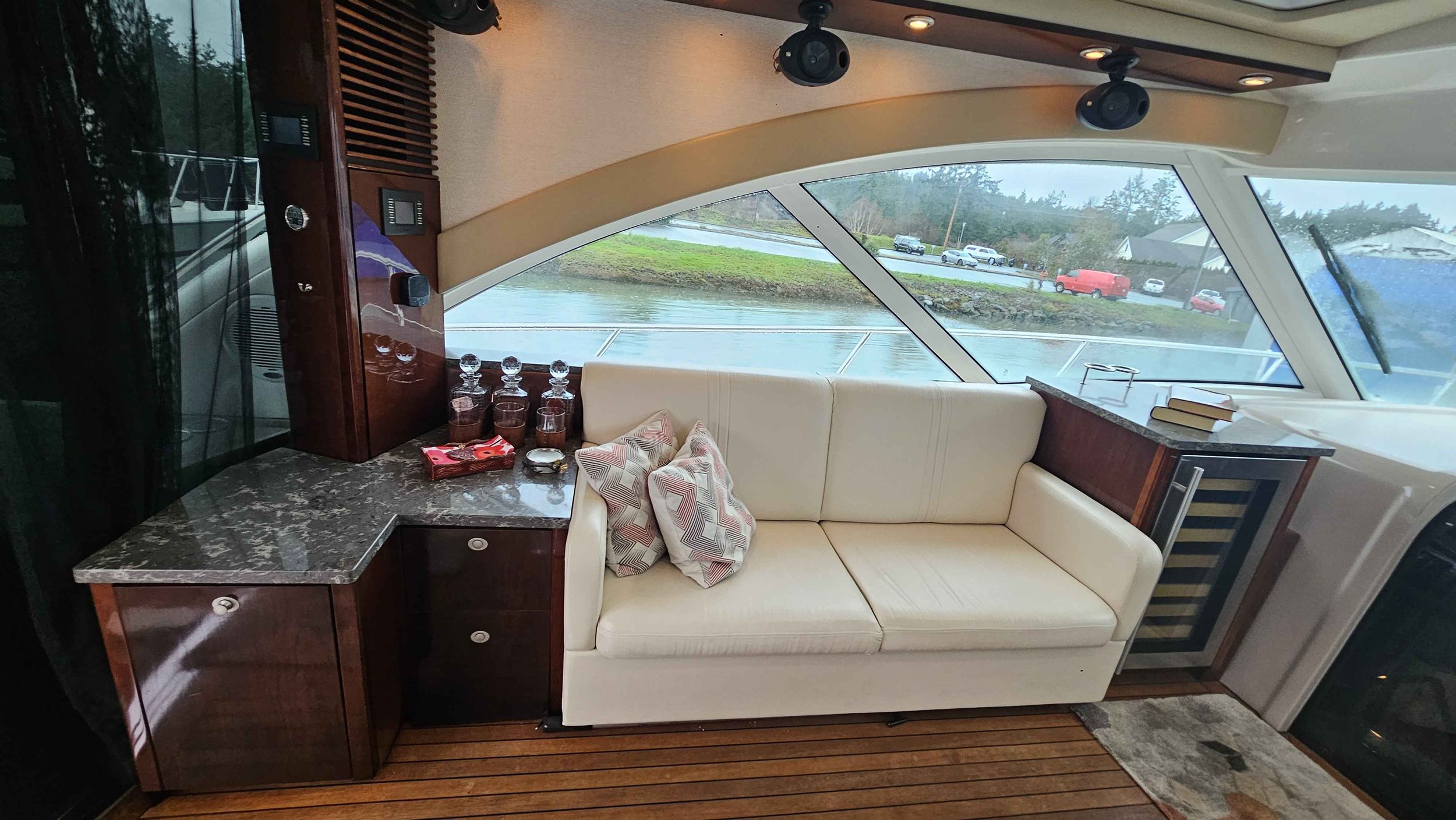 Interior of a yacht with a cream sofa, a stone-top table, books, and decorative glasses, overlooking waters and parked cars outside through large windows.