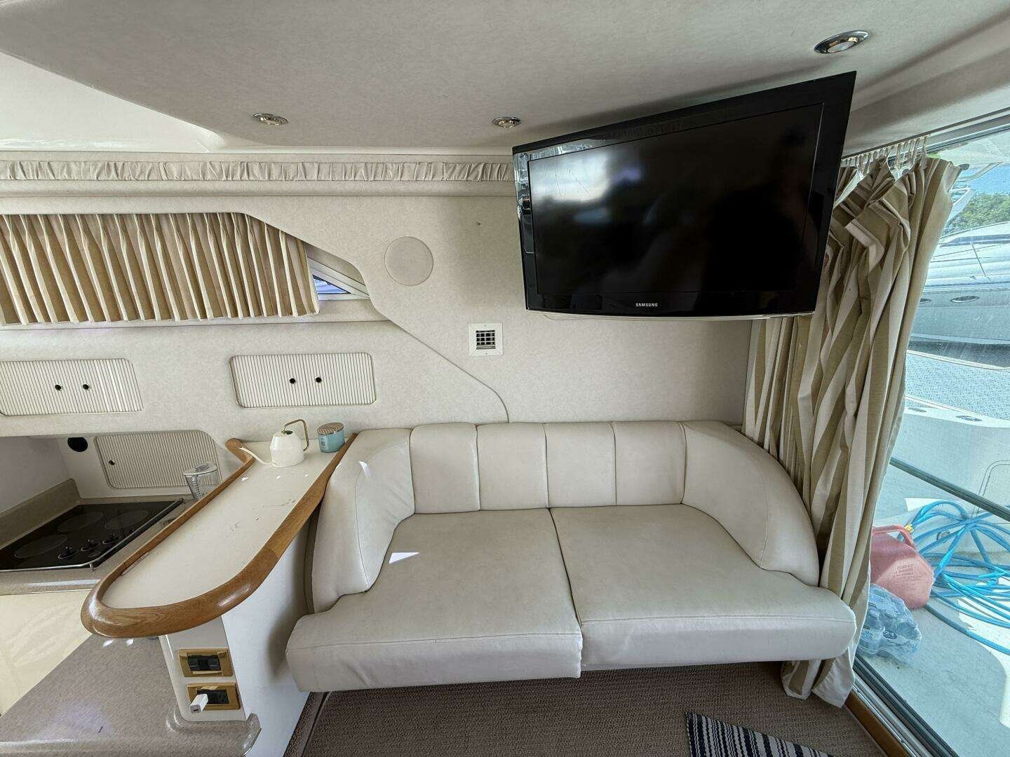 Interior of a luxury yacht with a corner white leather sofa, a mounted flat-screen TV, and a window with beige striped curtains.