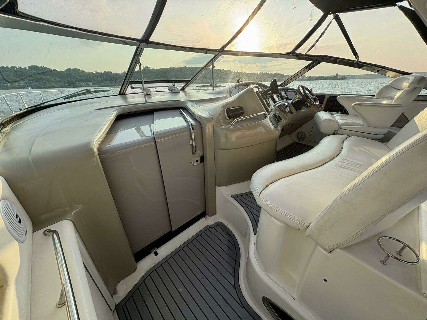 Helm view of a boat's cockpit with white leather seats, steering wheel, and navigation controls, overlooking a body of water during sunset. Sea Ray 460 Sundancer.
