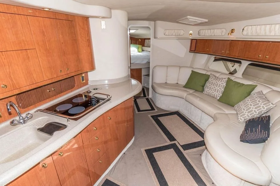 Interior of a boat with a curved white leather sofa, green and patterned pillows, wooden upper and lower cabinets, a small kitchen with a sink and electric stove, and a bedroom visible through a doorway.
