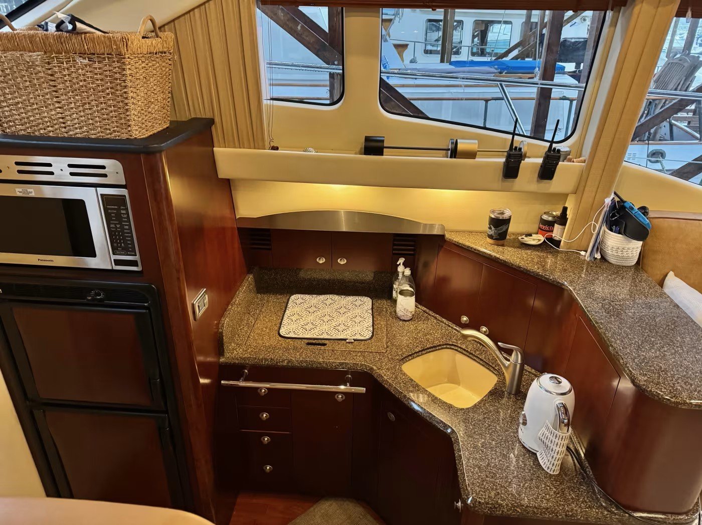 A yacht's kitchen with brown wood cabinets, granite countertops, small sink, microwave, and various small appliances and items on the counter, with a large window showing boats outside.