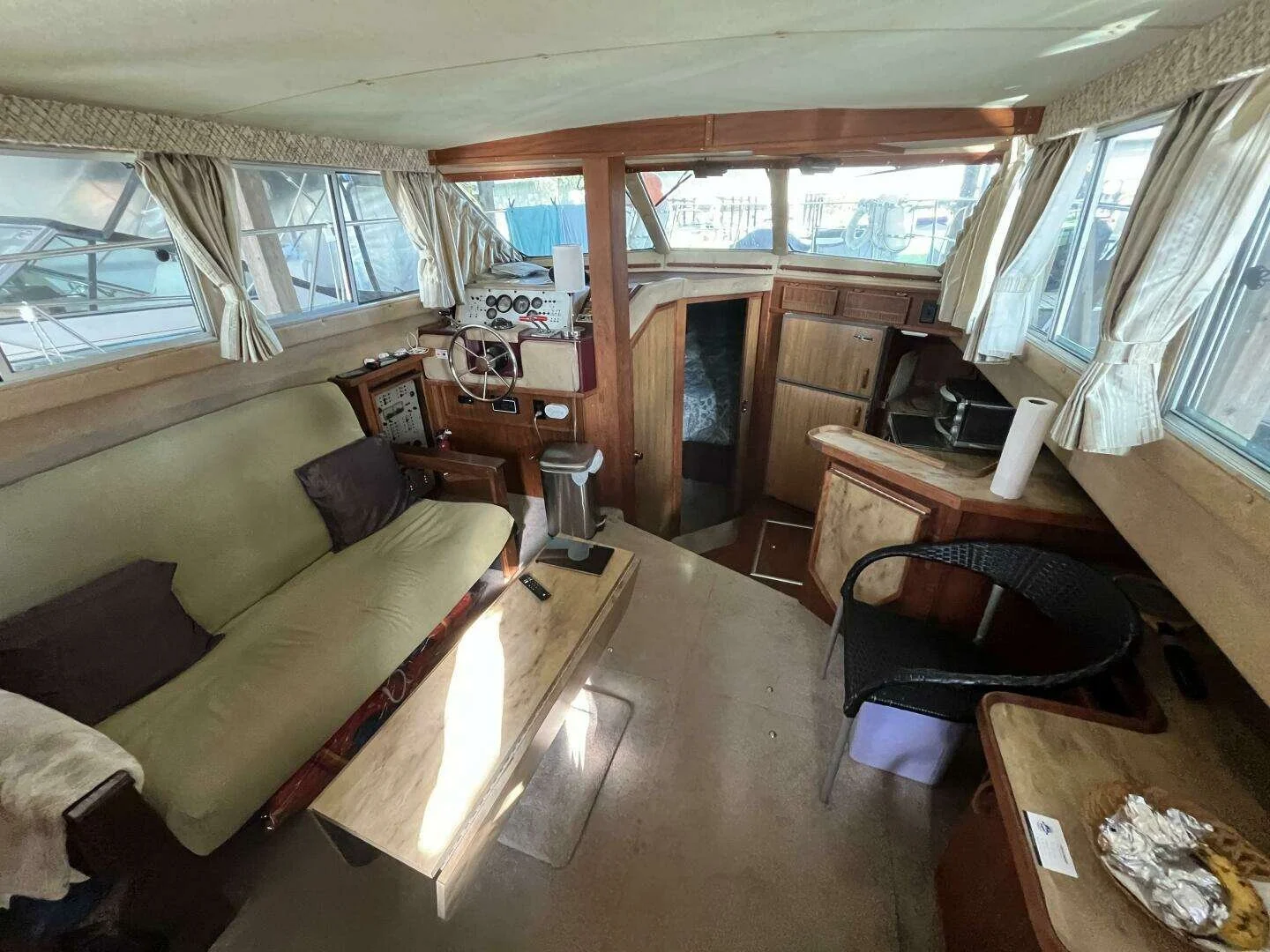 Interior view of a boat cabin with a green sofa, wooden cabinetry, windows with curtains, a small table, a chair, and navigation equipment.