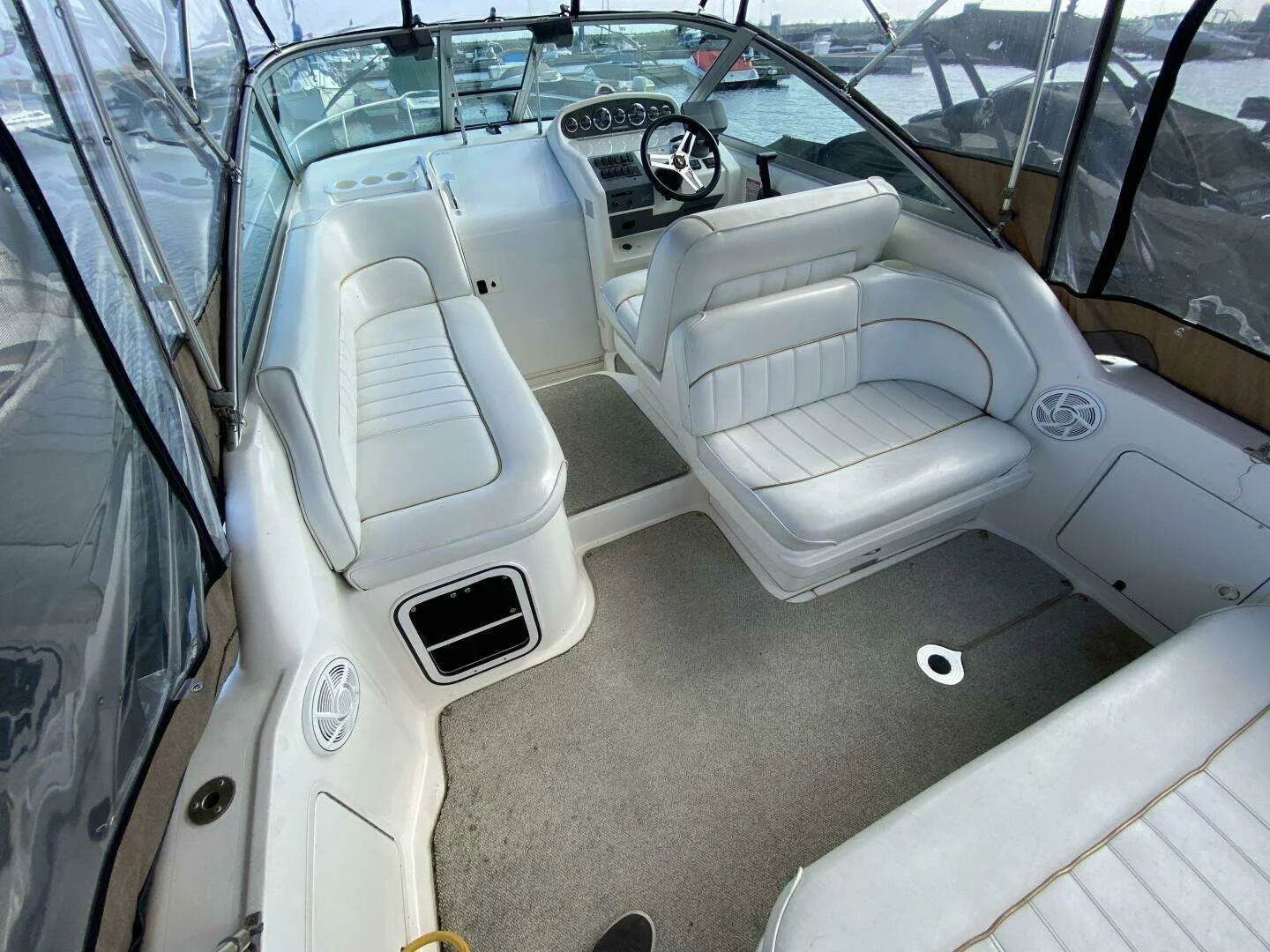 Cockpit view of a boat with white cushioned seating, a steering wheel, and a helm control panel. The cabin is enclosed with large windows and is docked at a marina. Sea Ray 270 Sundancer.