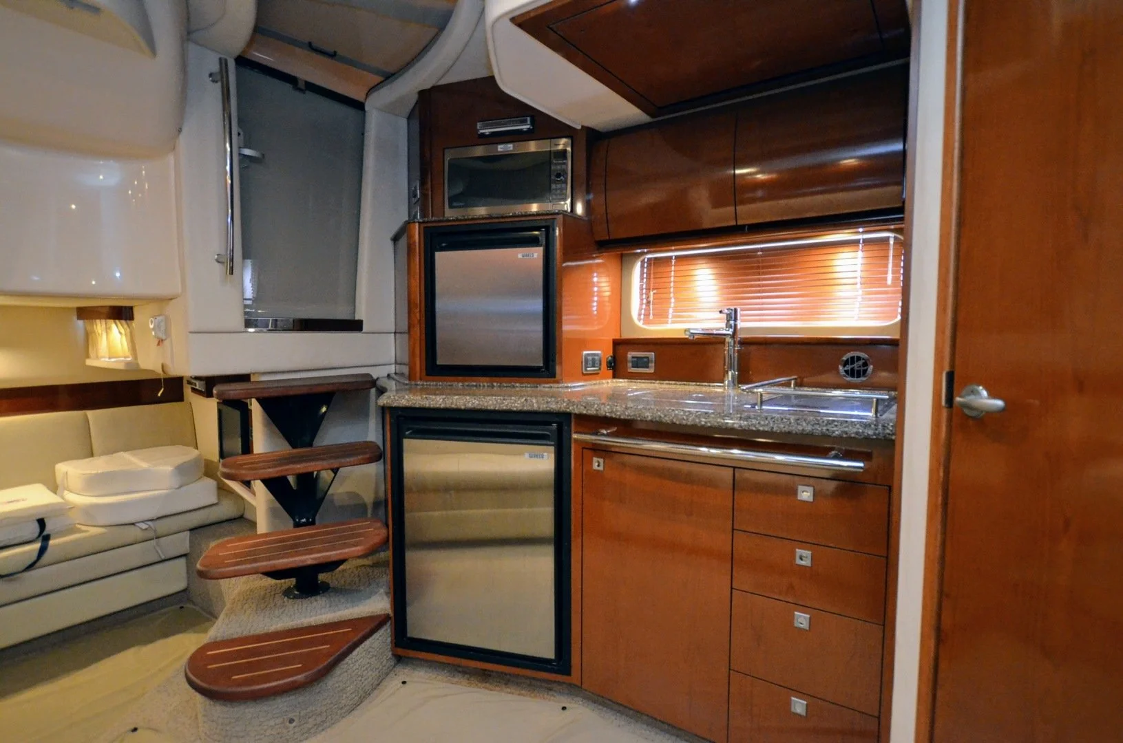 Interior of a yacht kitchen with wooden cabinetry, a granite countertop, a small sink, and a window with blinds. There are built-in appliances including a microwave and a refrigerator, along with wooden stairs leading to a higher level and a seating 