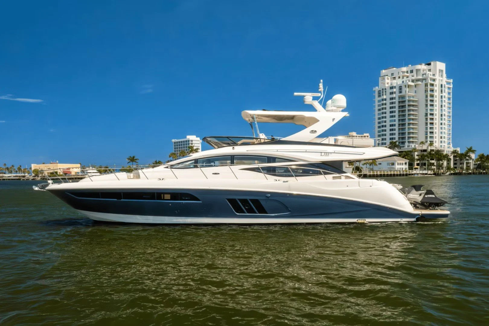 Luxury yacht cruising on water with city buildings and palm trees in background under clear blue sky.