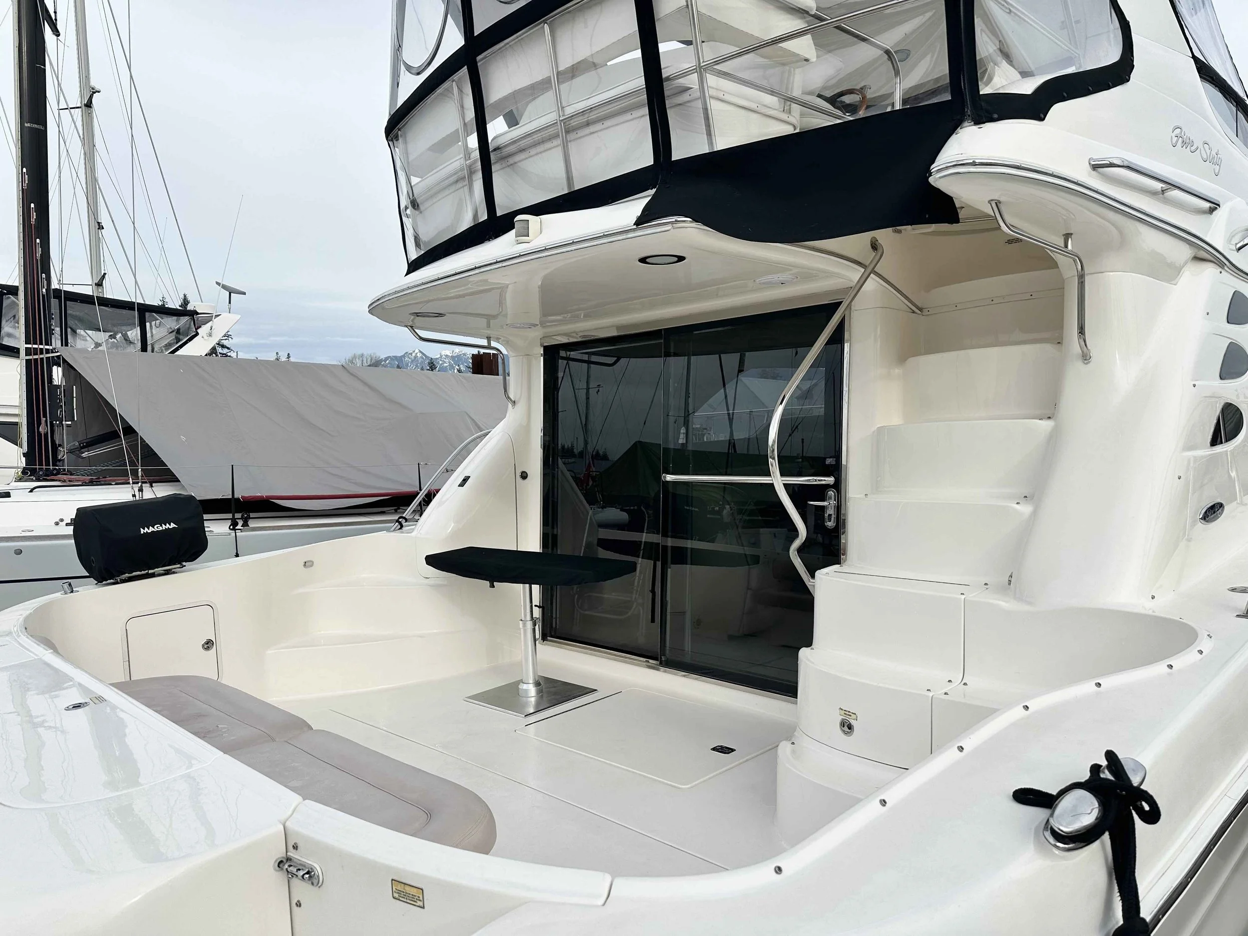 A white yacht docked at a marina with a small seating area on the deck, a sliding glass door leading inside, and various other boats in the background.