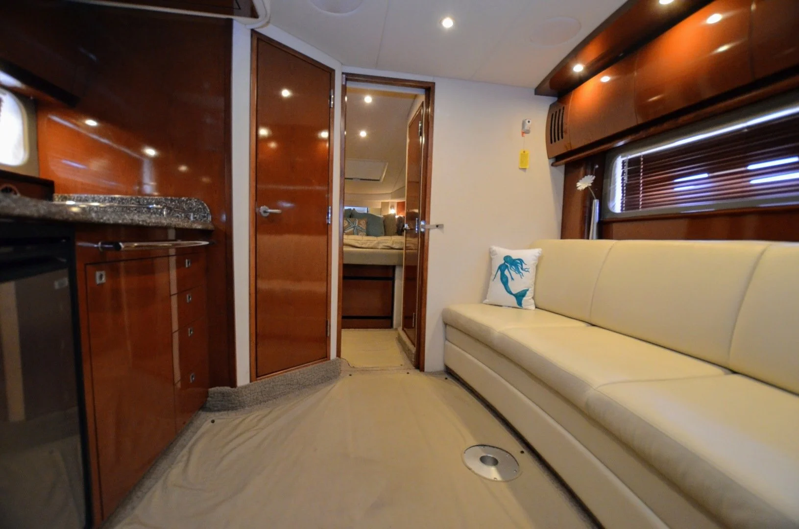 Interior of a boat or yacht with a beige sofa, a cushion with a blue mermaid design, a window with closed blinds, and wooden cabinetry, leading to a bedroom with a bed and pillows.