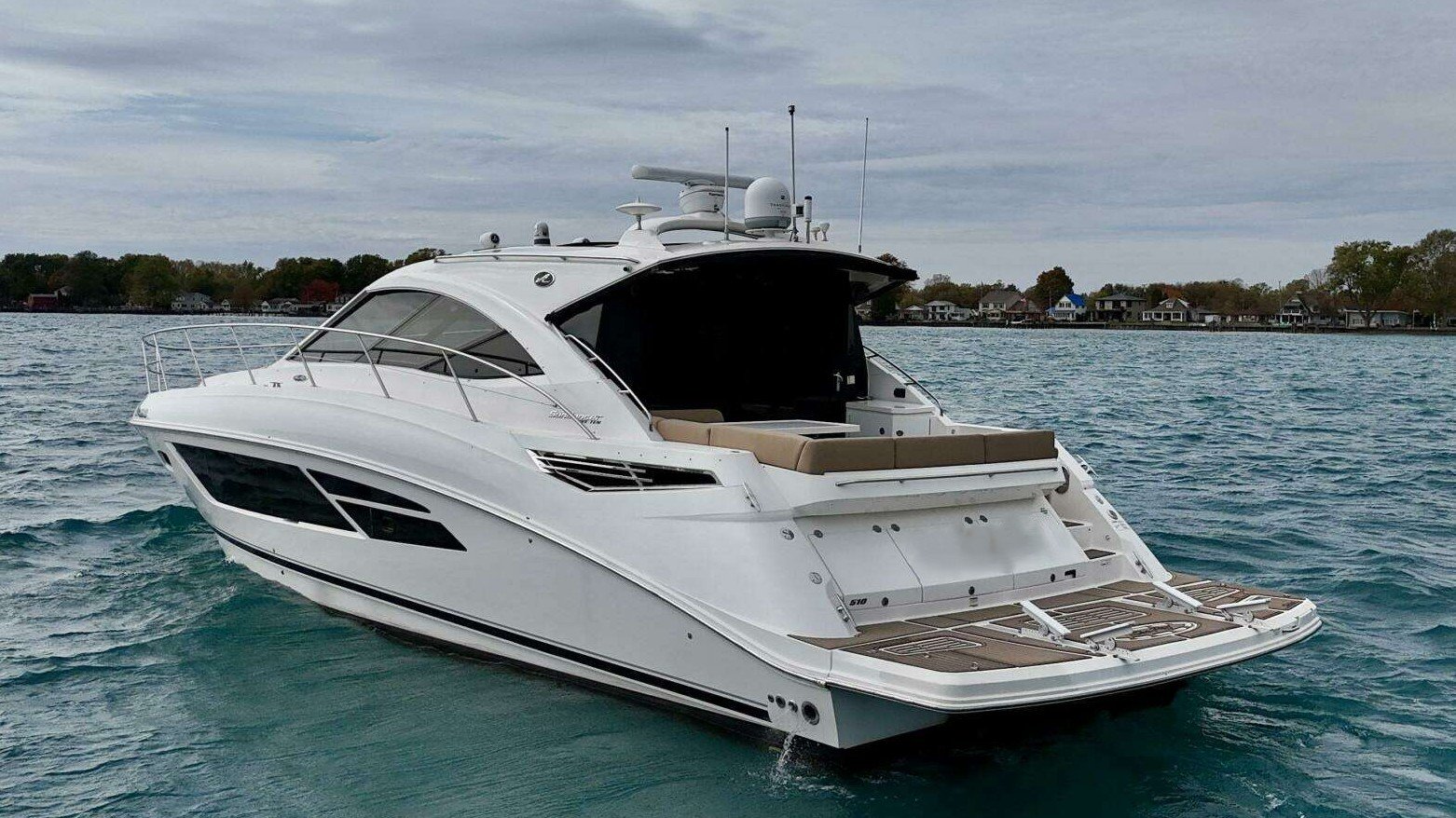 A white luxury motor yacht on the water with a spacious deck and a covered seating area, near a shoreline with houses and trees.