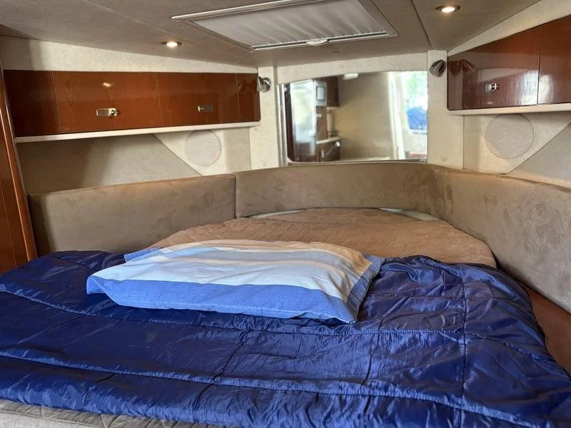 Interior view of a camper or RV bedroom with a large bed covered in blue and beige bedding, beige cushioned headboard, wooden cabinets, and a mirror.