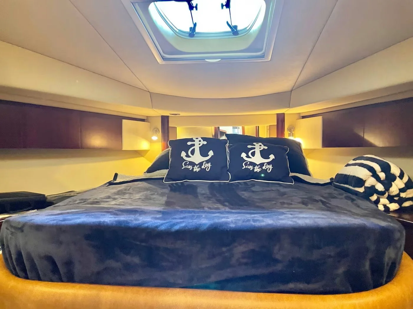 Bedroom with a round bed covered in a velvet dark blue blanket, two decorative pillows with anchor designs and the phrase 'Seas the Day,' and a striped black and white blanket on the side. A skylight window in the ceiling on this Sea Ray yacht.