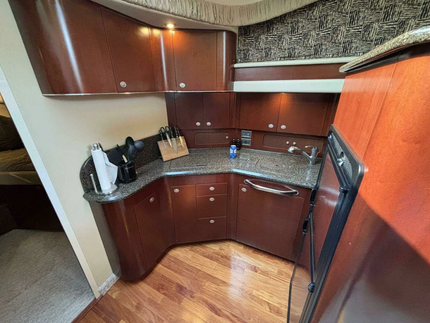 A Sea Ray boat kitchen galley area with dark wood cabinets, a granite countertop, a sink, several knives in a wooden block, paper towel and utensils, and a refrigerator on the right.