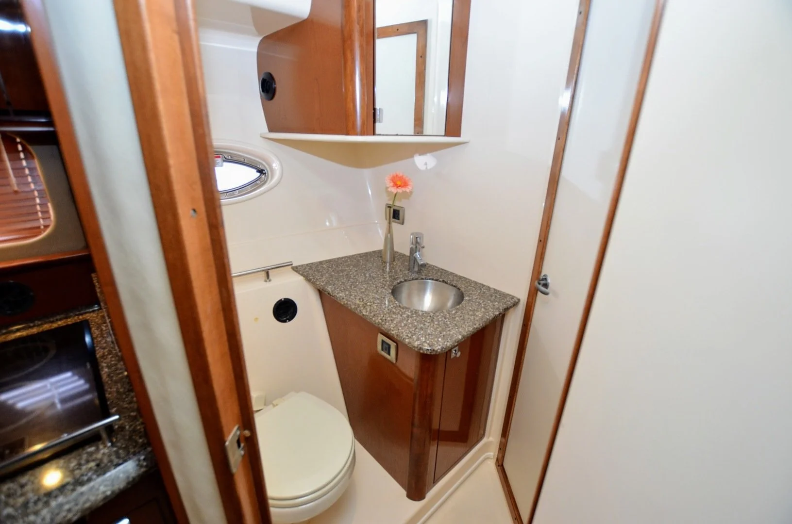 Interior of a yacht's bathroom with a small granite sink, a wooden cabinet below, a small potted flower, a mirror above, and a tiny oval window.