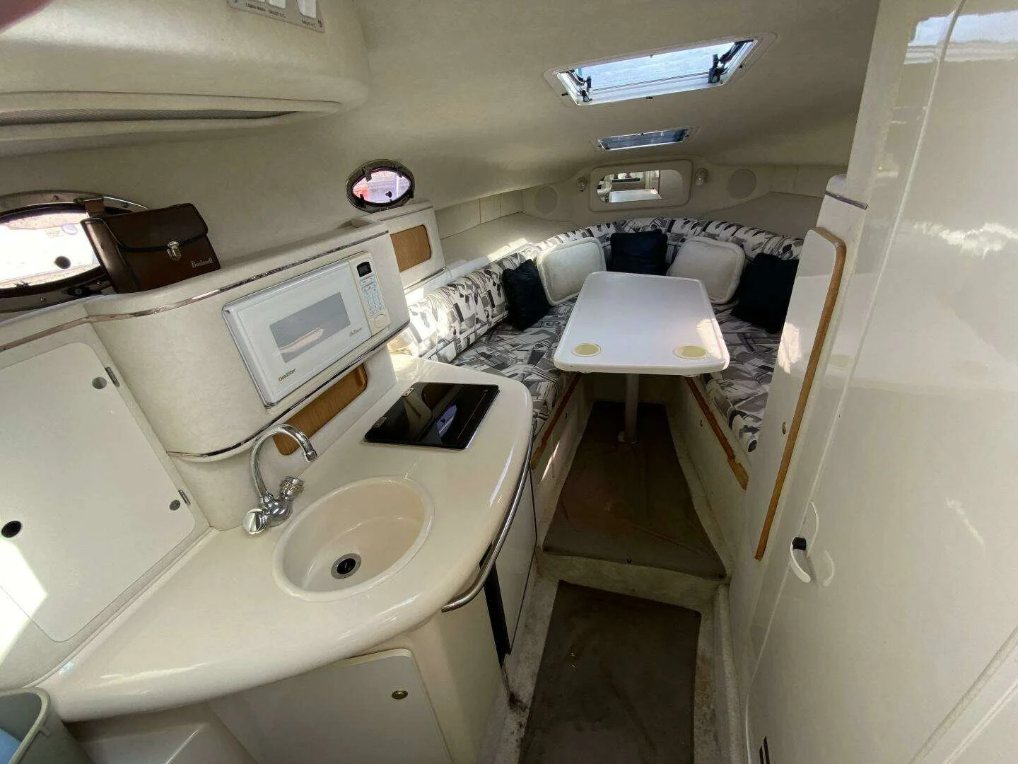 Interior of a small yacht with a small kitchen area on the left, including a microwave, sink, and stovetop, and a seating area with a table and cushioned benches on the right, under small windows. Sea Ray 270 Sundancer boat.