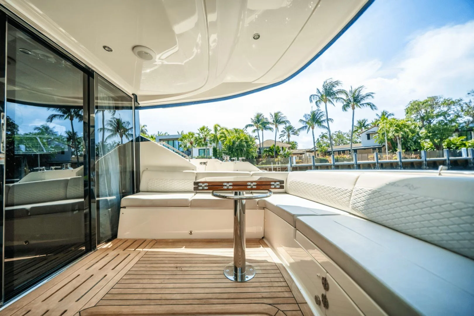 View of the deck area on a luxury yacht with white cushioned seating, a small round table, and sliding glass doors, with palm trees and houses visible in the background. Sea Ray boat.