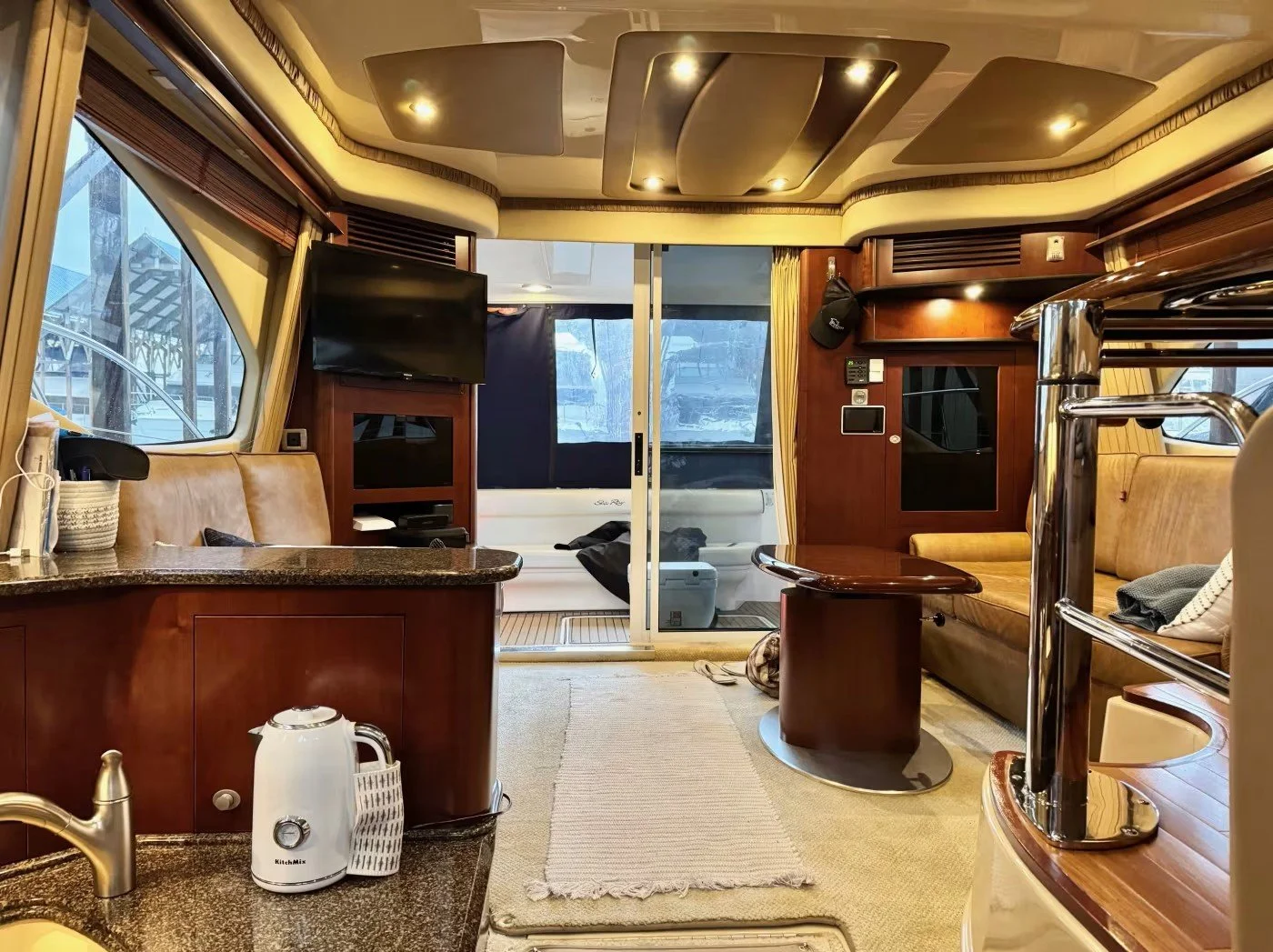 Interior of a yacht living area with leather seating, wooden cabinetry, a television, sliding glass door leading outside, a small table, and a kitchen counter with a kettle.