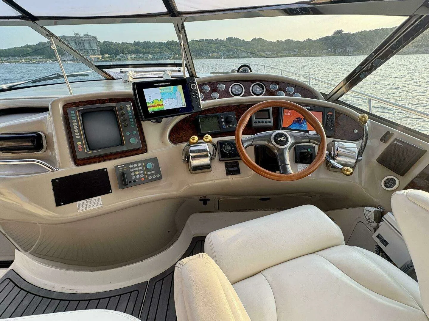 View of a boat's helm with navigation and control instruments, a steering wheel, and a partial view of water and shoreline outside. Sunny summer evening. Sea Ray 460 Sundancer. 