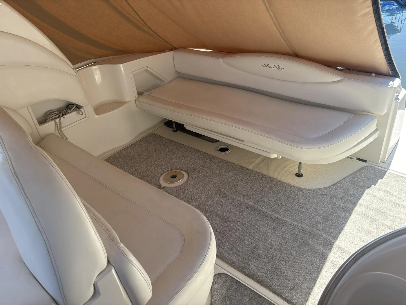 Interior of a boat with white cushioned seating, a carpeted floor, and a wooden ceiling, with a small hole and a circular object on the floor.